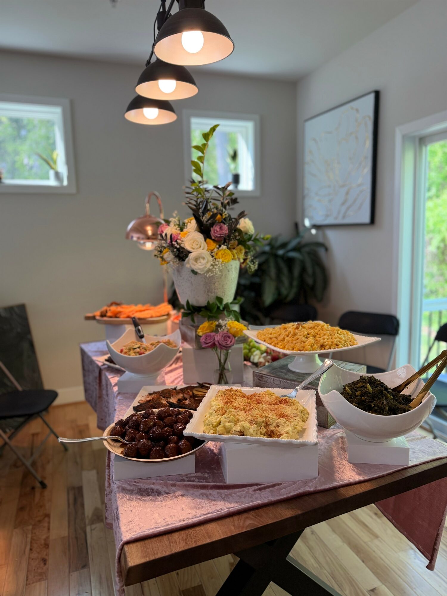 Dining table with a flower arrangement and various dishes, illuminated by ceiling lights, with windows and a whiteboard in the background.