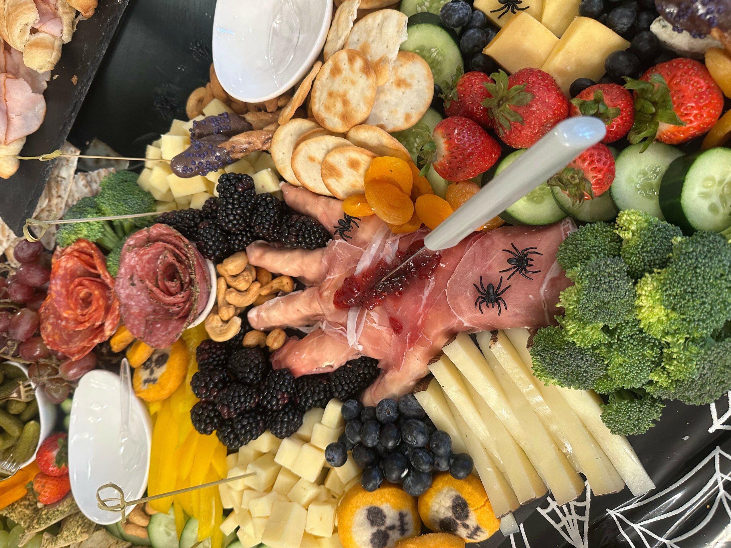 Assorted fruits, vegetables, cheese, and snacks arranged on a platter with a small spoon, including strawberries, broccoli, cheese slices, and grapes.