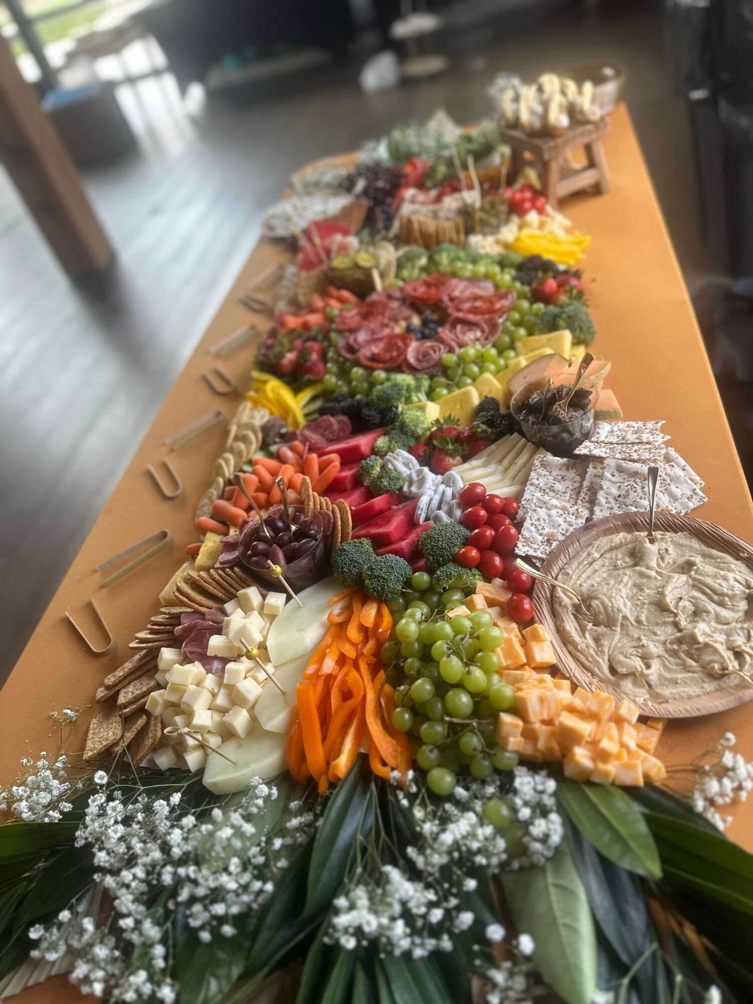 Colorful assortment of cheeses, fruits, and crackers arranged on a long wooden table, with flowers and greenery in the foreground.