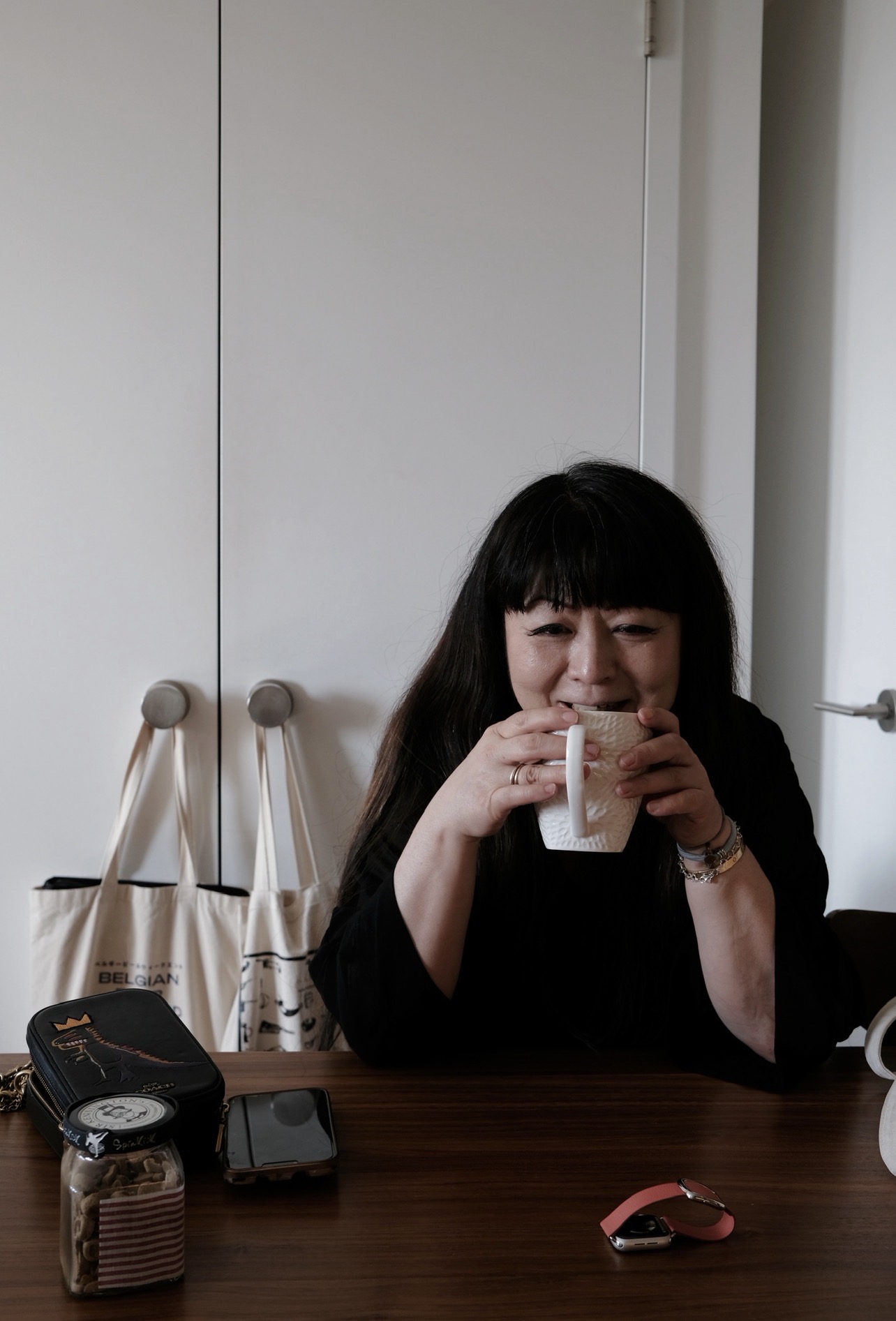 Woman with long dark hair sitting at a table, drinking from a glass, smiling, with a white wall behind her.