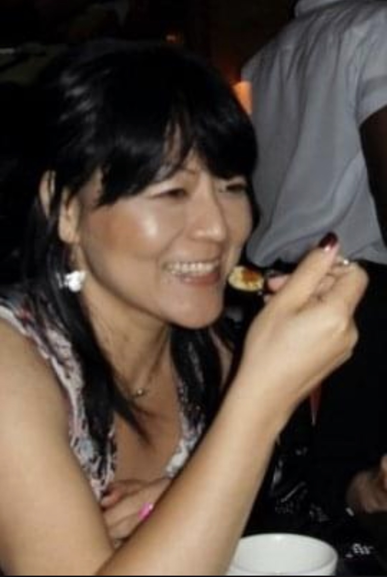 Woman smiling while eating, holding a spoon, with dark hair and earrings, in a social setting.