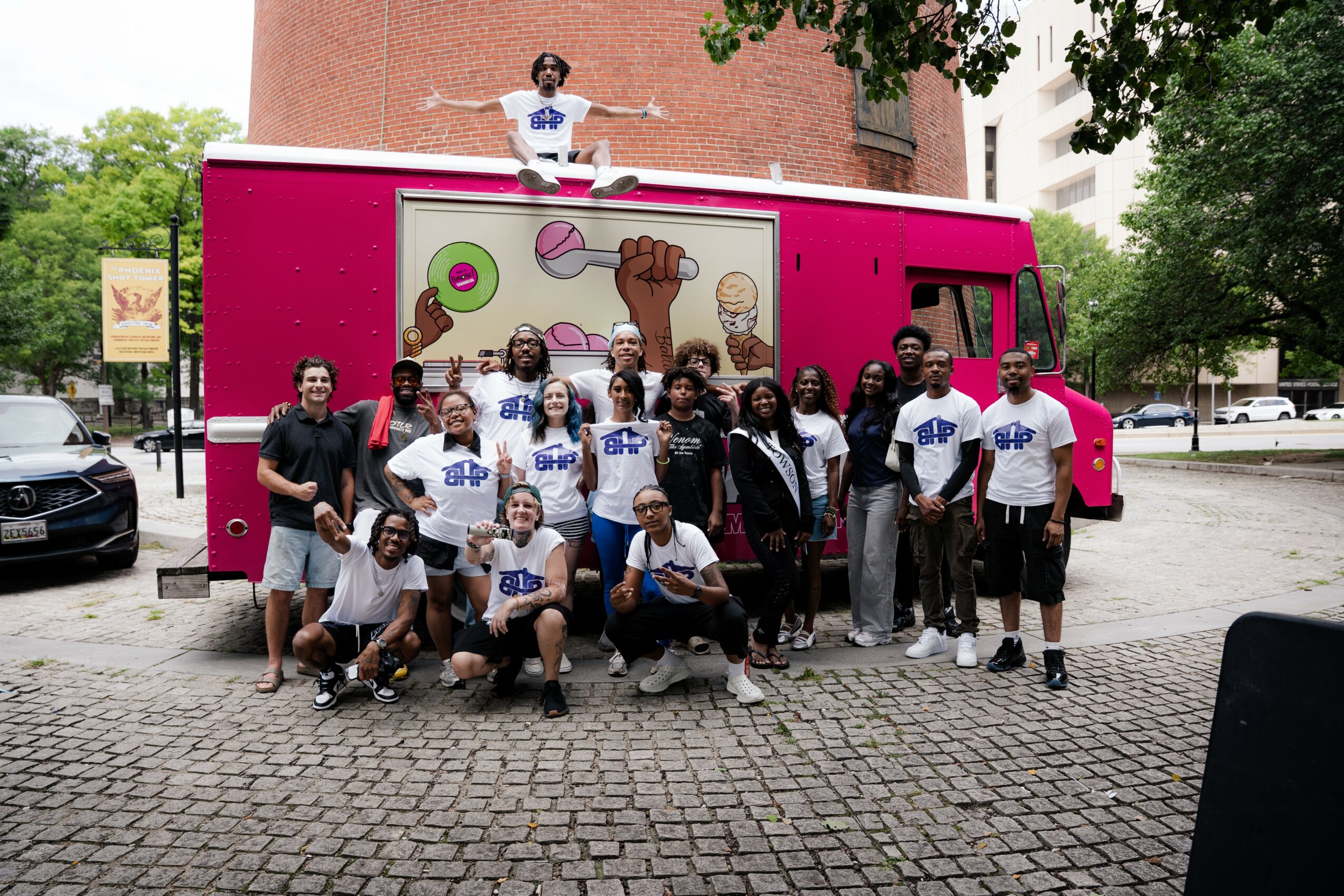 Group of people posing in front of a pink mobile food truck with a colorful mural, some standing and some kneeling, outdoors.