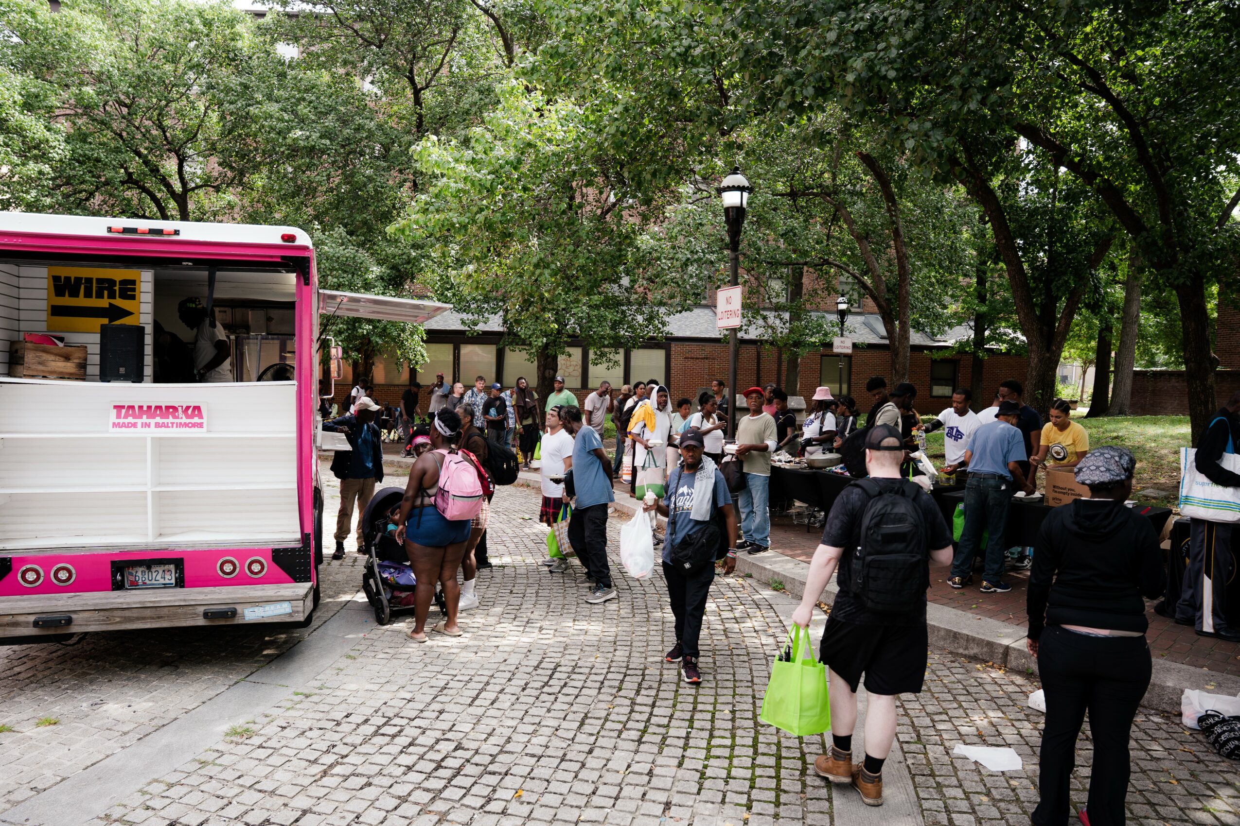People gather in a park with trees, some standing, others sitting, near a pink food truck, with a cobblestone ground.