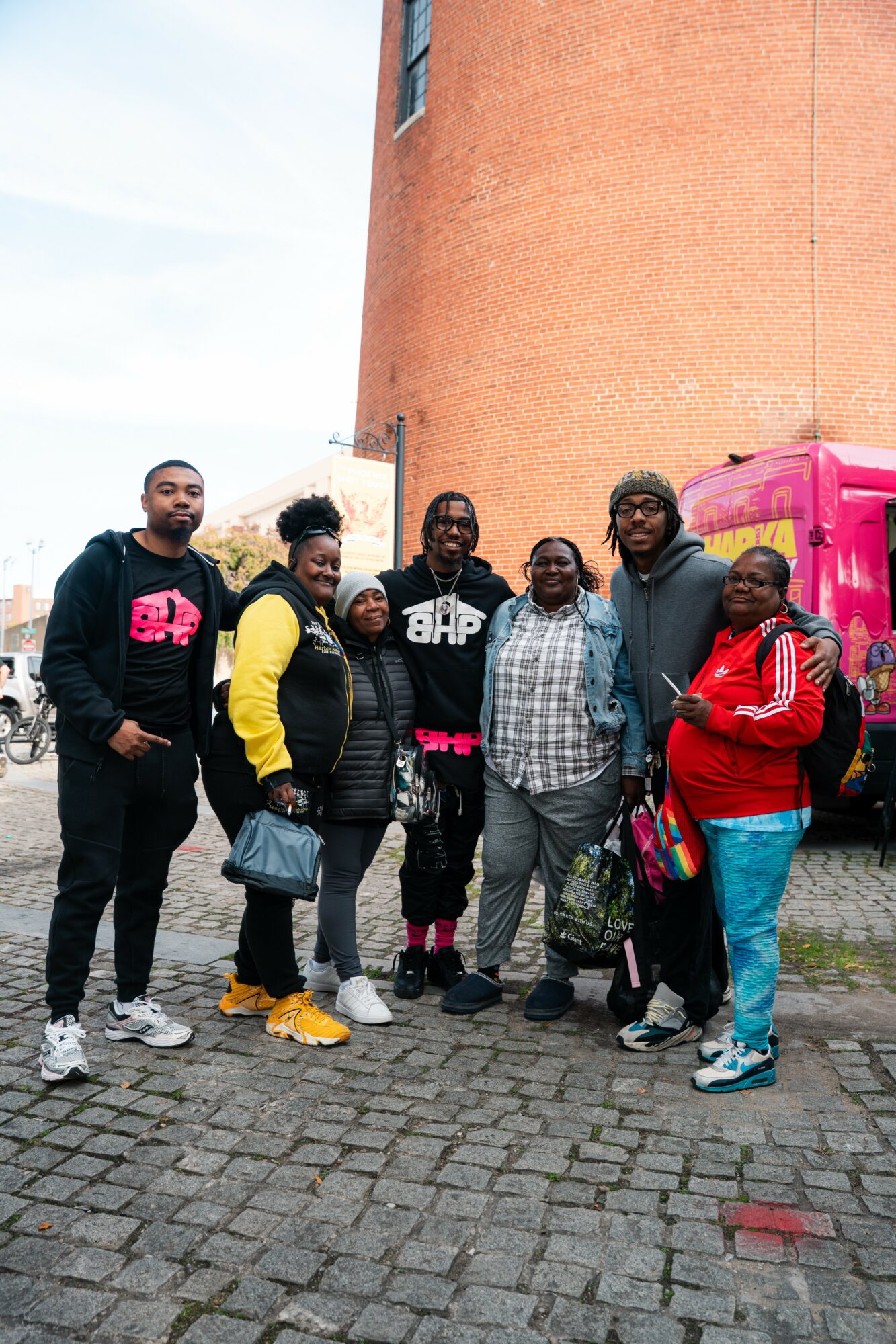 Group of six people standing on cobblestone street in front of a brick building and pink food truck.