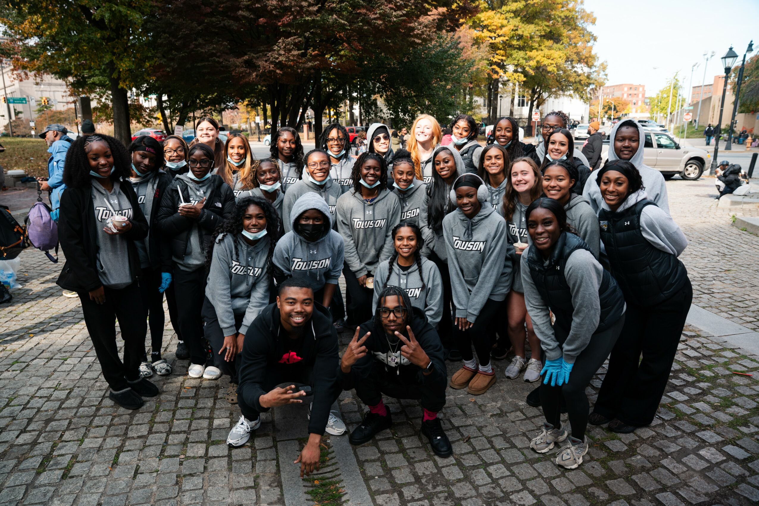 Group of young people outdoors on cobblestone street, some wearing hoodies, trees with autumn leaves in background.