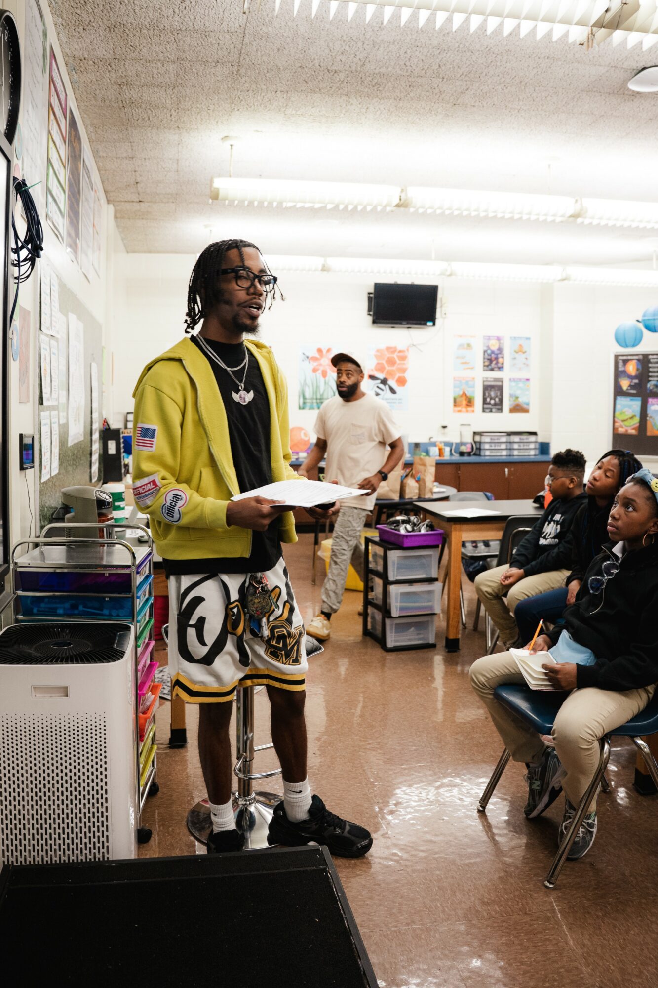 Person standing in front of a classroom, holding a paper, with students seated and listening attentively.