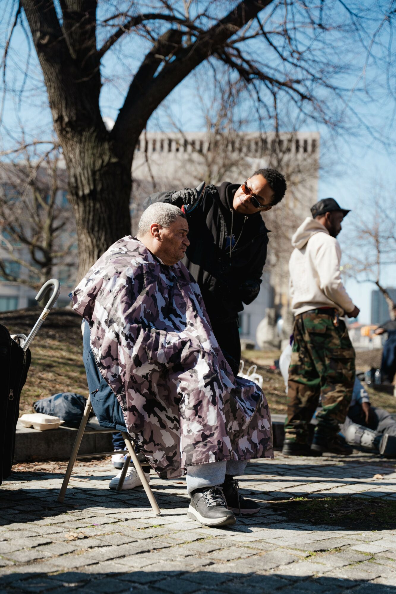 Two men, one seated and one standing, converse outdoors near a large tree with a building in the background.