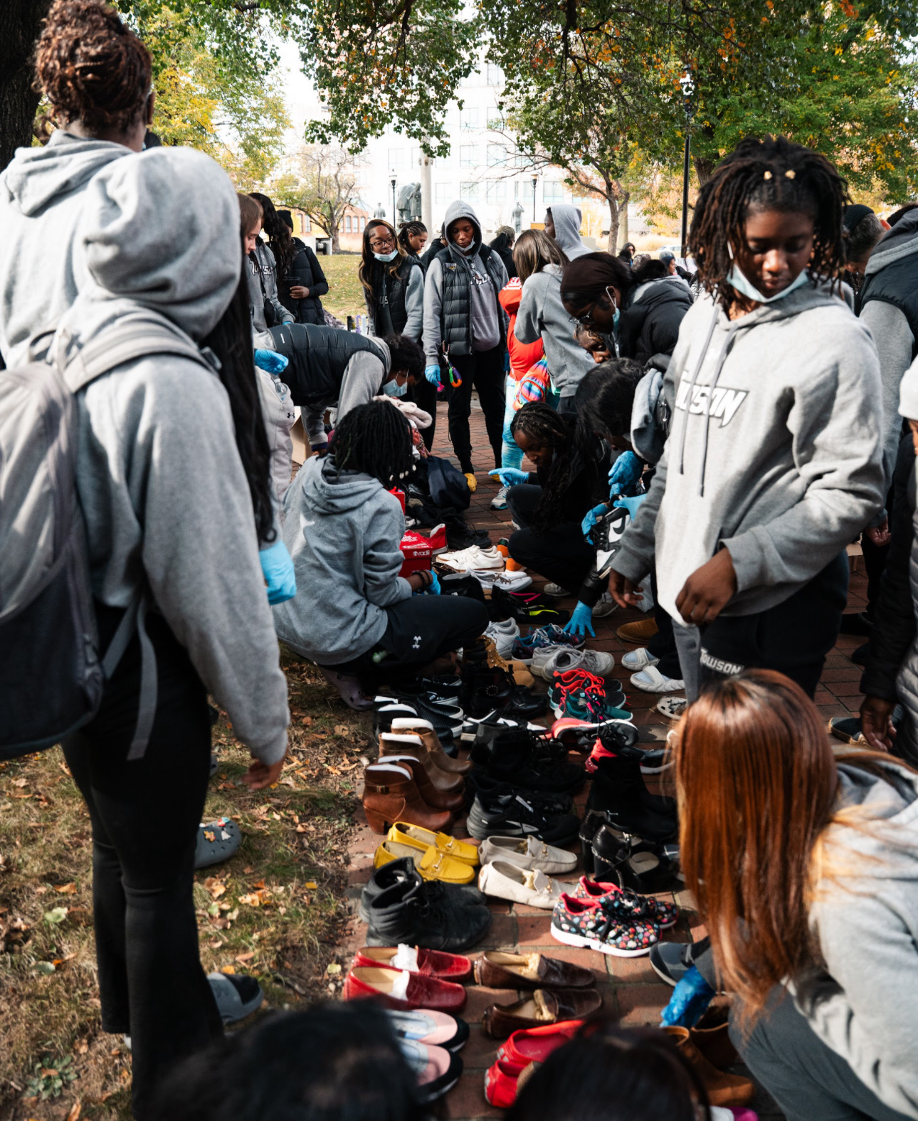 Group of people outdoors browsing shoes on the ground, trees and sunlight in the background.