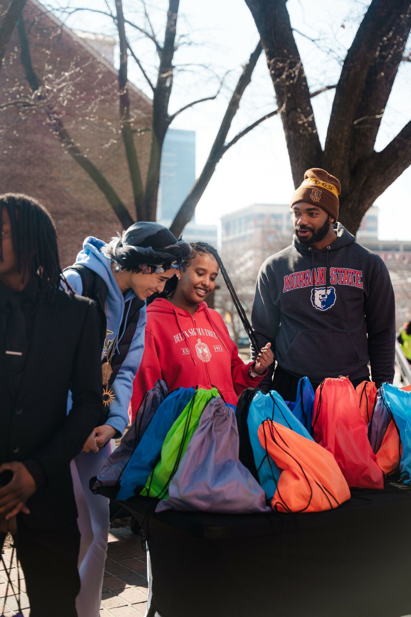 Group of people outdoors with trees and buildings, standing near a table with colorful backpacks, smiling and talking.