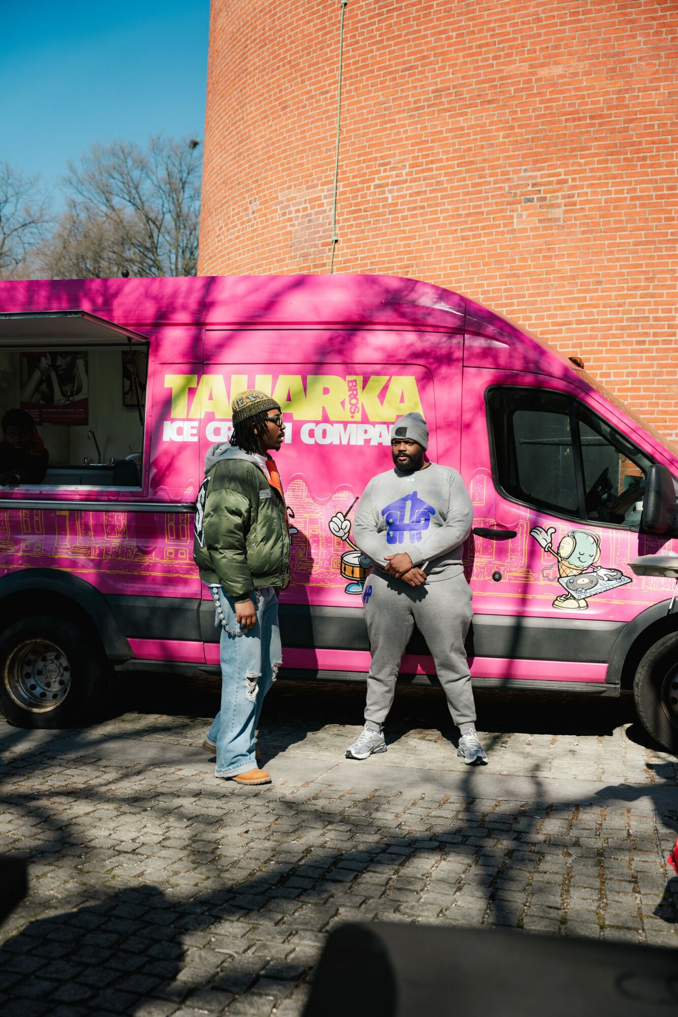 Two men stand in front of a pink van with text, one wearing a green jacket and jeans, the other in a gray hoodie and gray pants, with a brick wall and trees in the background.