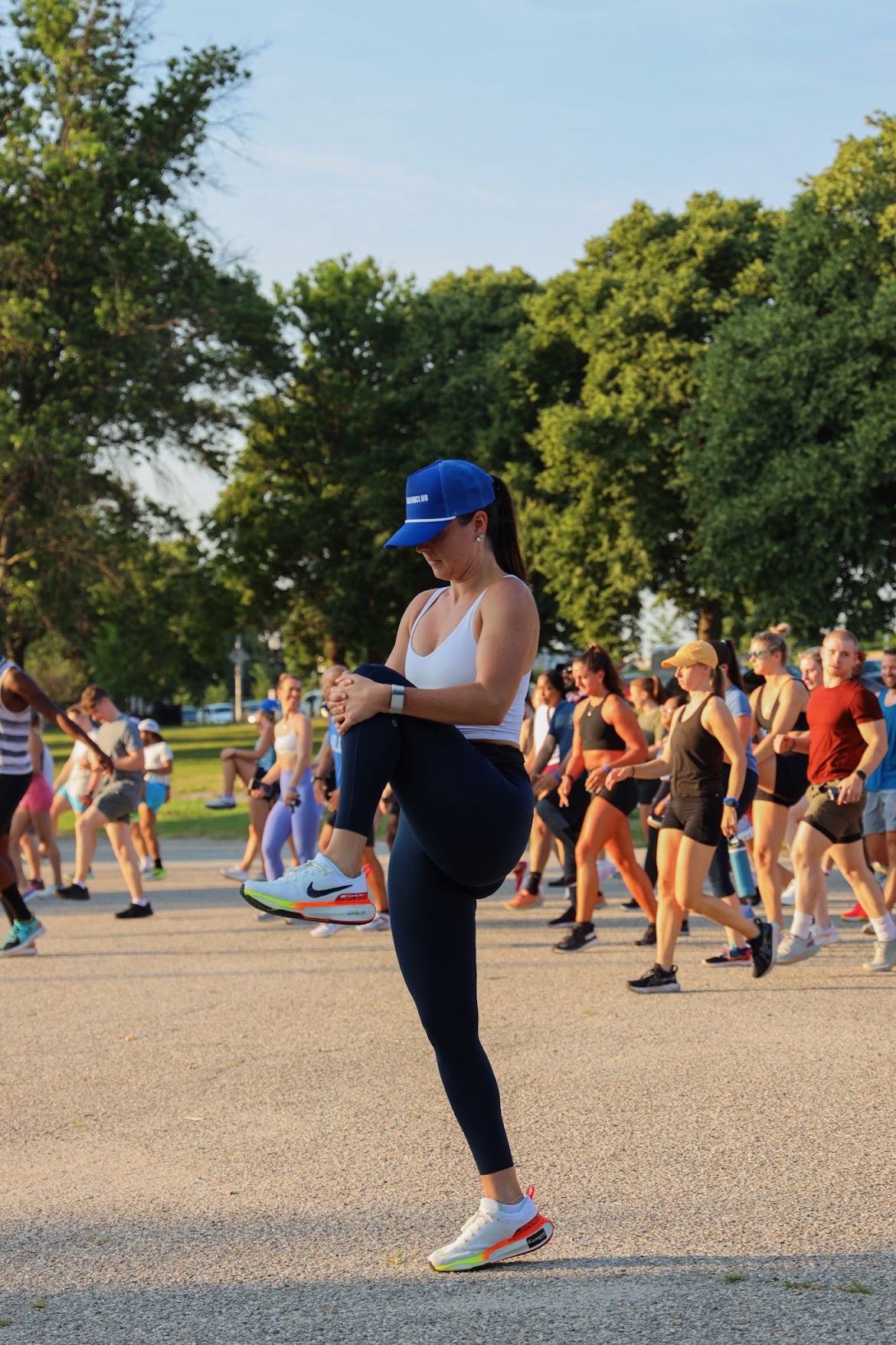Woman in white tank top, blue cap, and leggings standing outdoors among people, trees, and a clear sky.