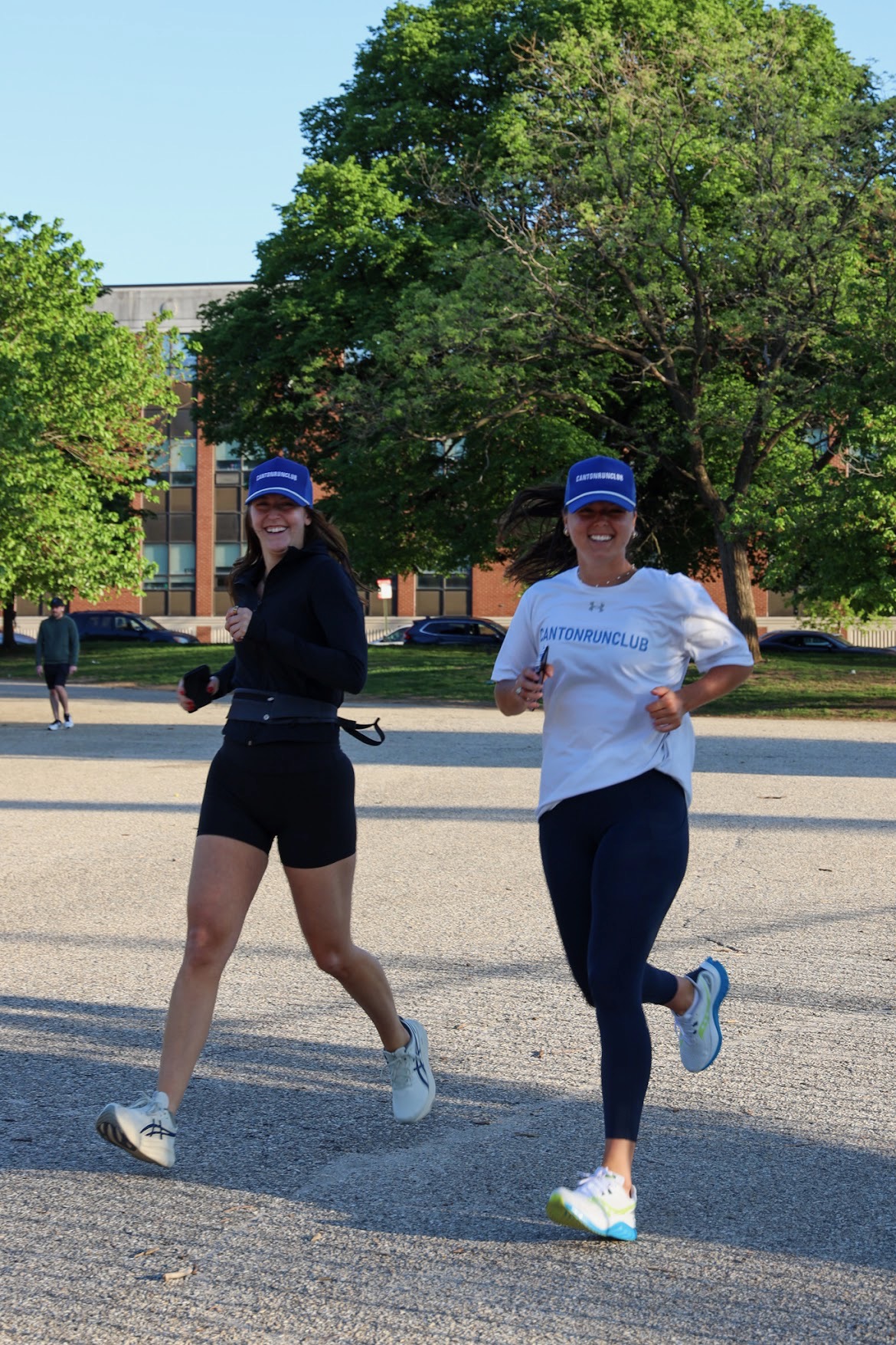 Two women jogging outdoors on a sunny day, wearing athletic clothing and caps, with trees and a building in the background.