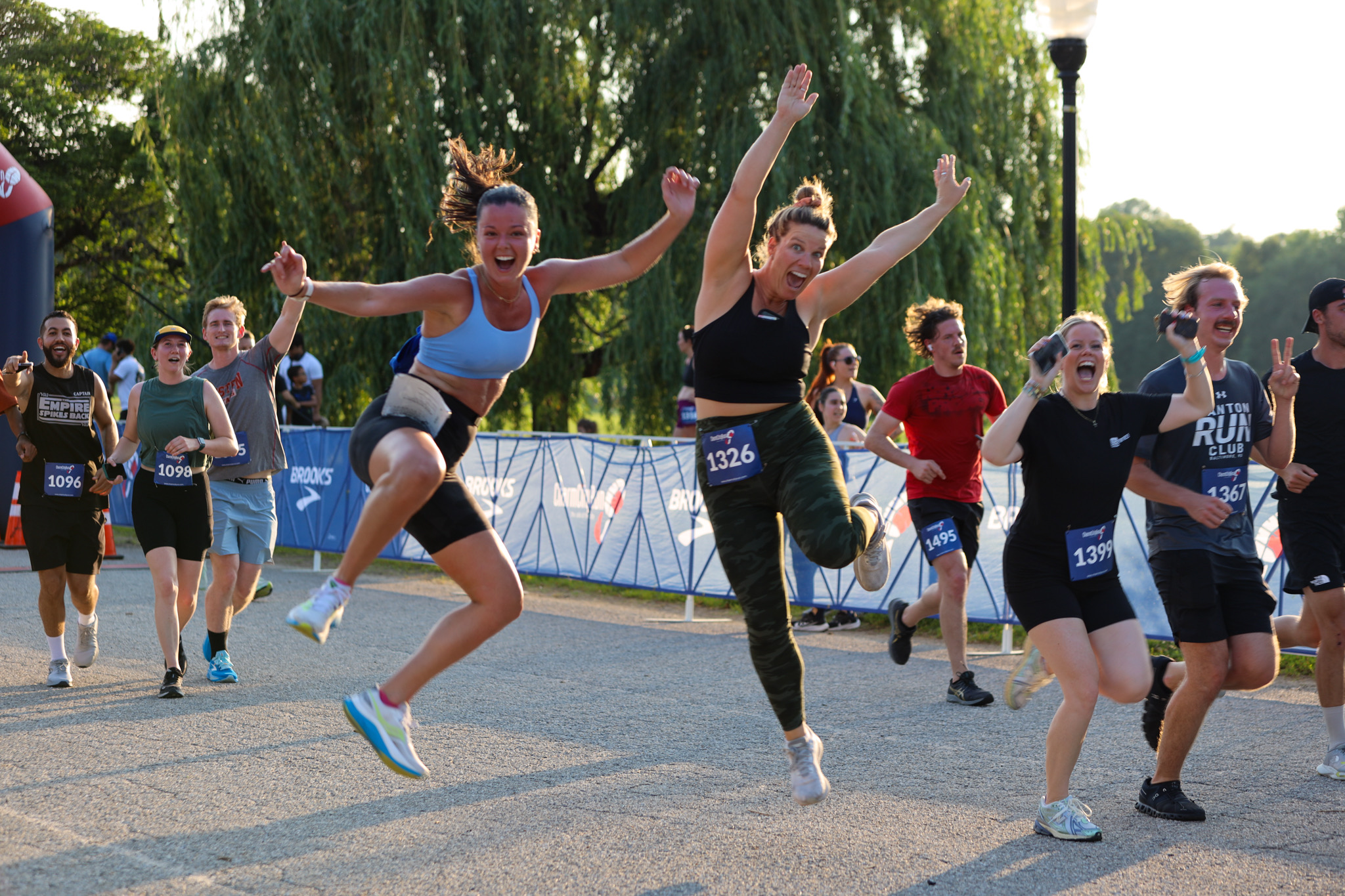 Group of people running in a race, some jumping and smiling outdoors with trees and a blue sky.