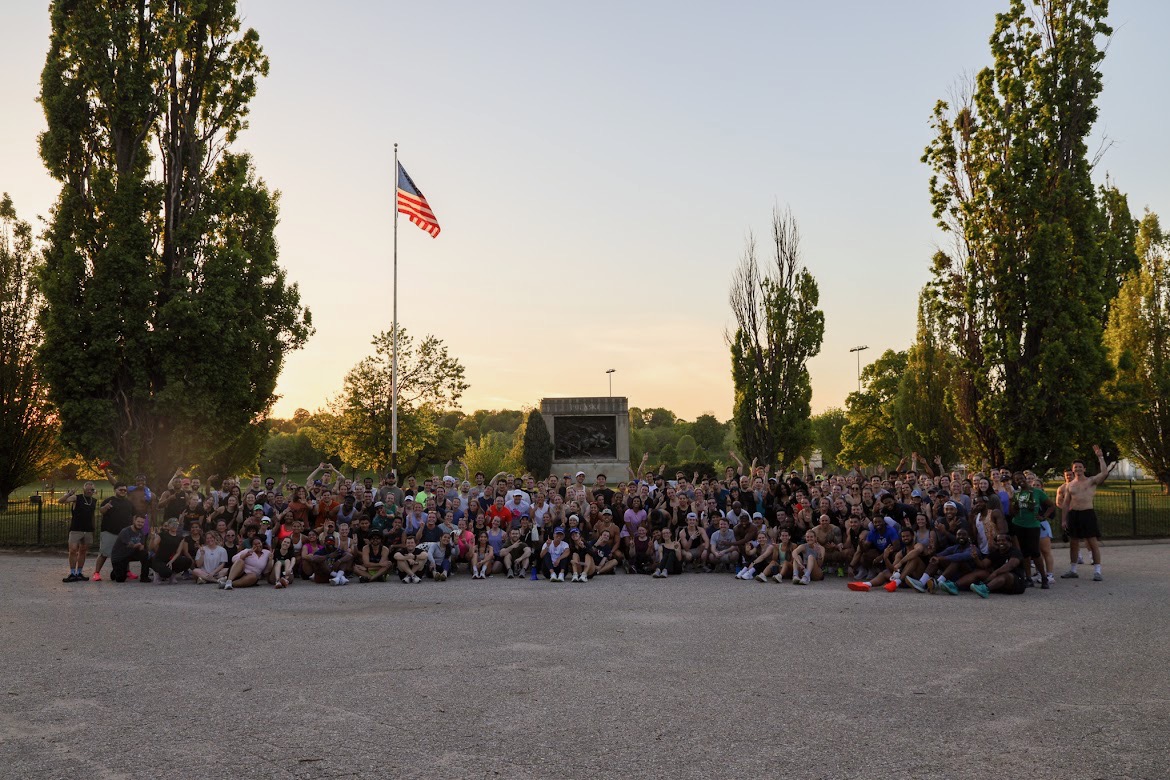Large group of people gathered outdoors in front of a monument, with trees and an American flag, during daytime.