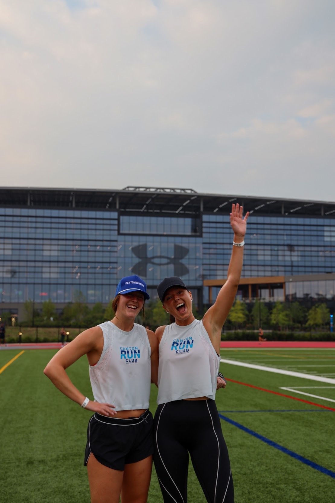 Two women in athletic clothing and caps standing on a sports field, smiling, with a large modern building in the background.
