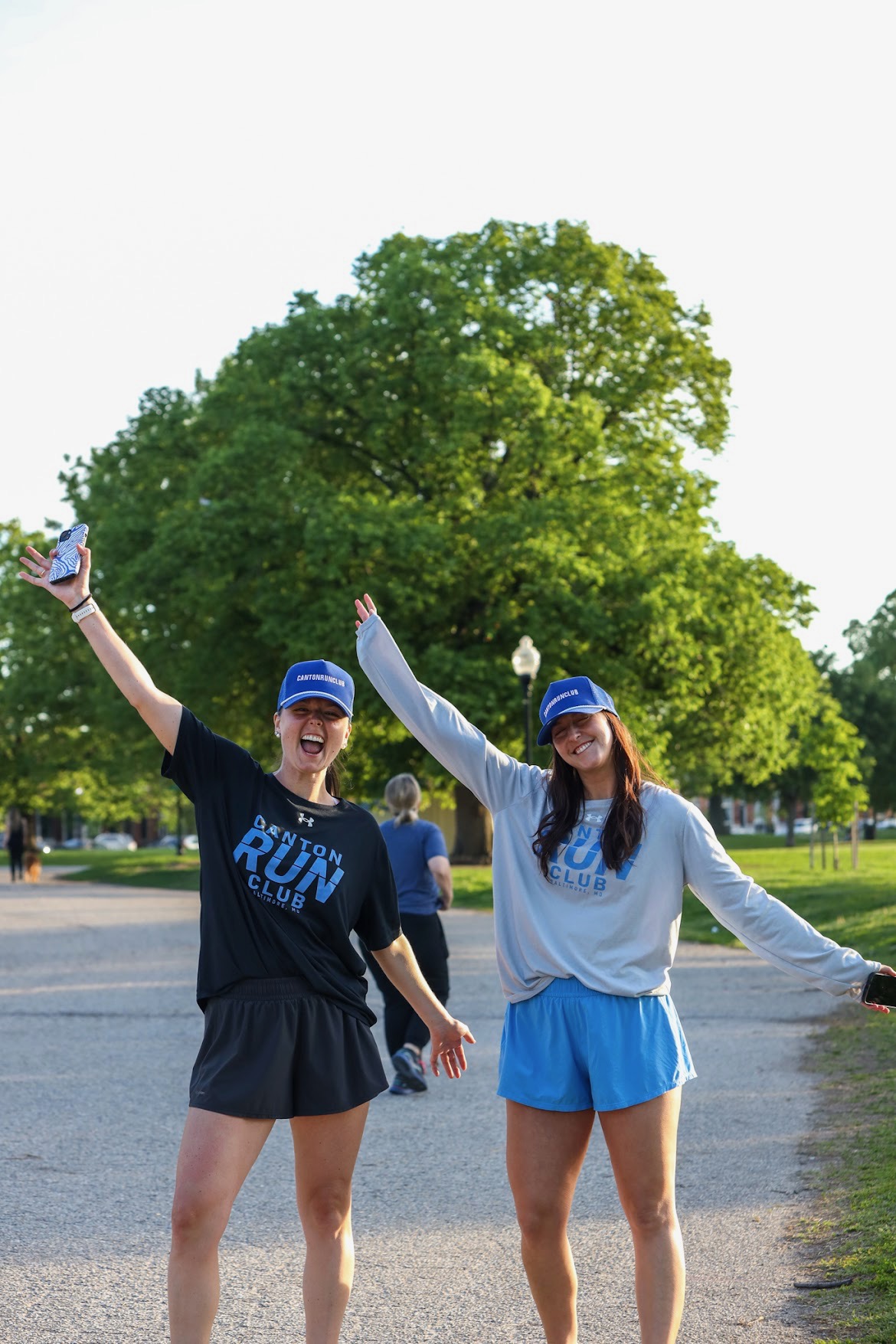 Two women in athletic clothing and caps smiling and raising their arms outdoors with trees in the background.
