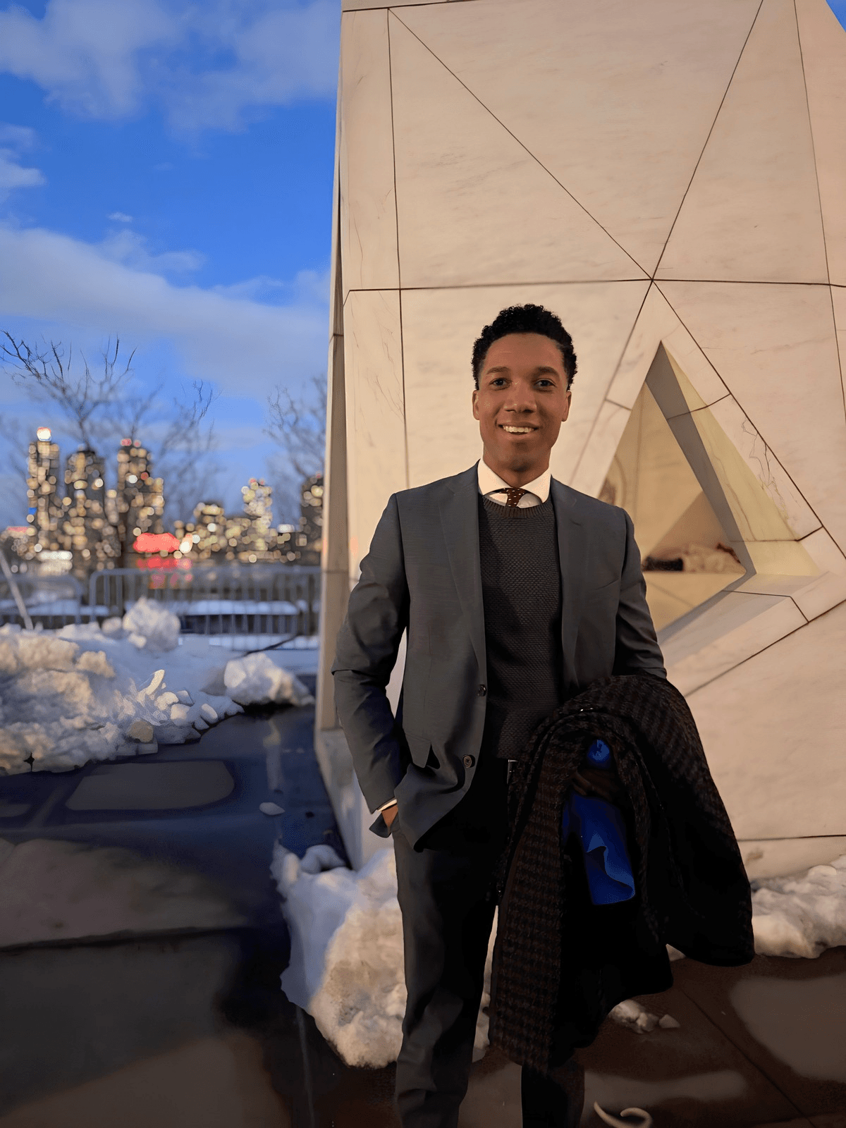 Young man in a suit standing outdoors near a modern building with snow and cityscape in the background.