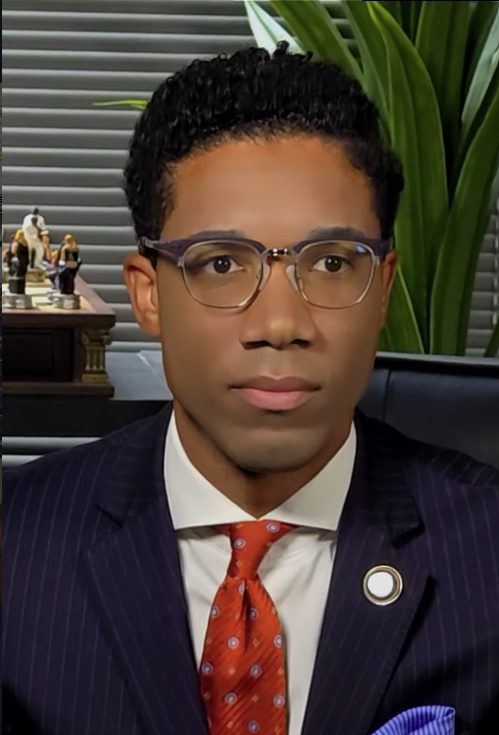 Young man with glasses in a suit and tie, sitting indoors with plants and decorative items behind him.
