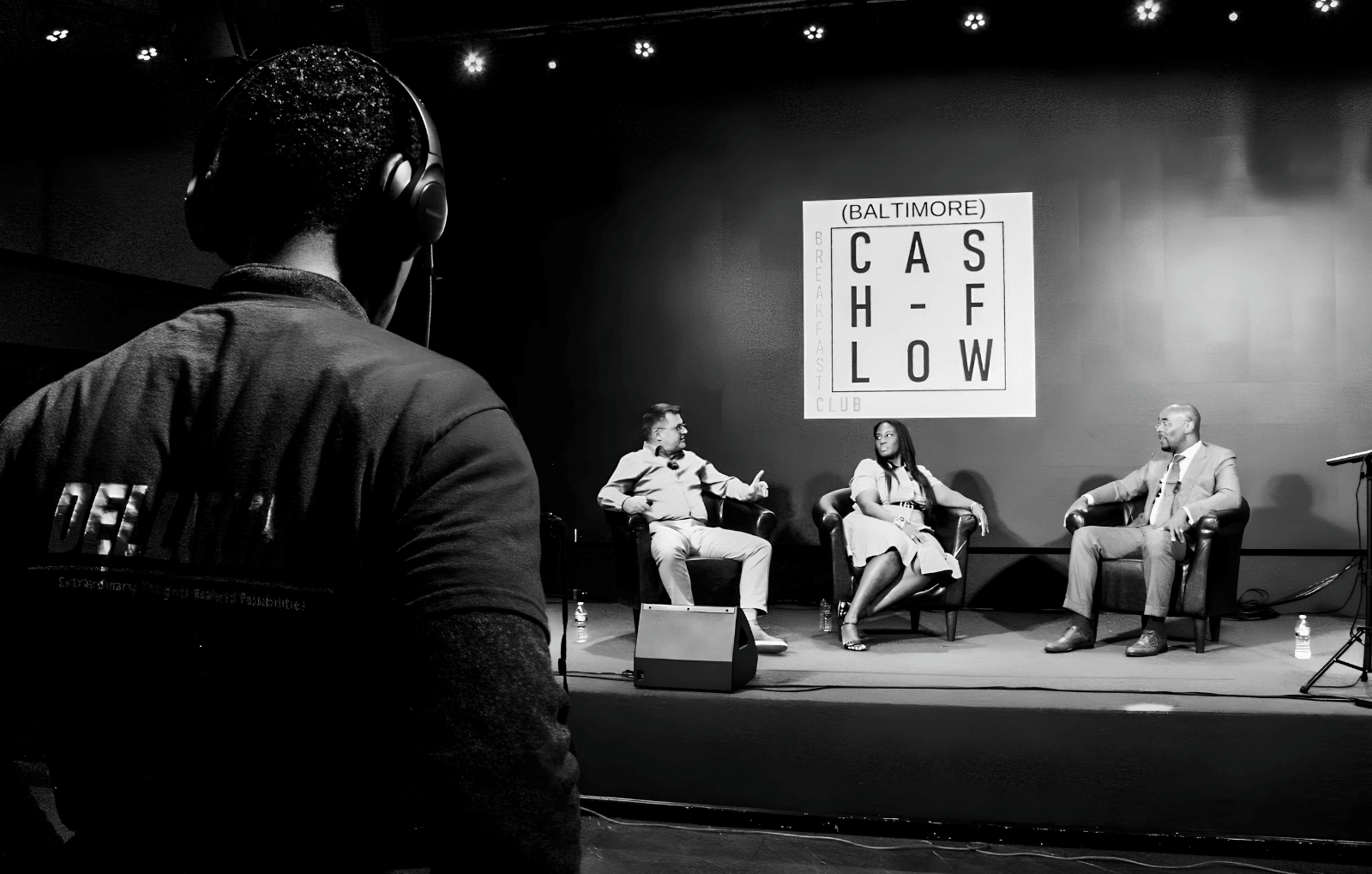 Person with headphones watching a panel discussion on stage with three people, and a sign reading 'Baltimore' and 'Chow' behind them.