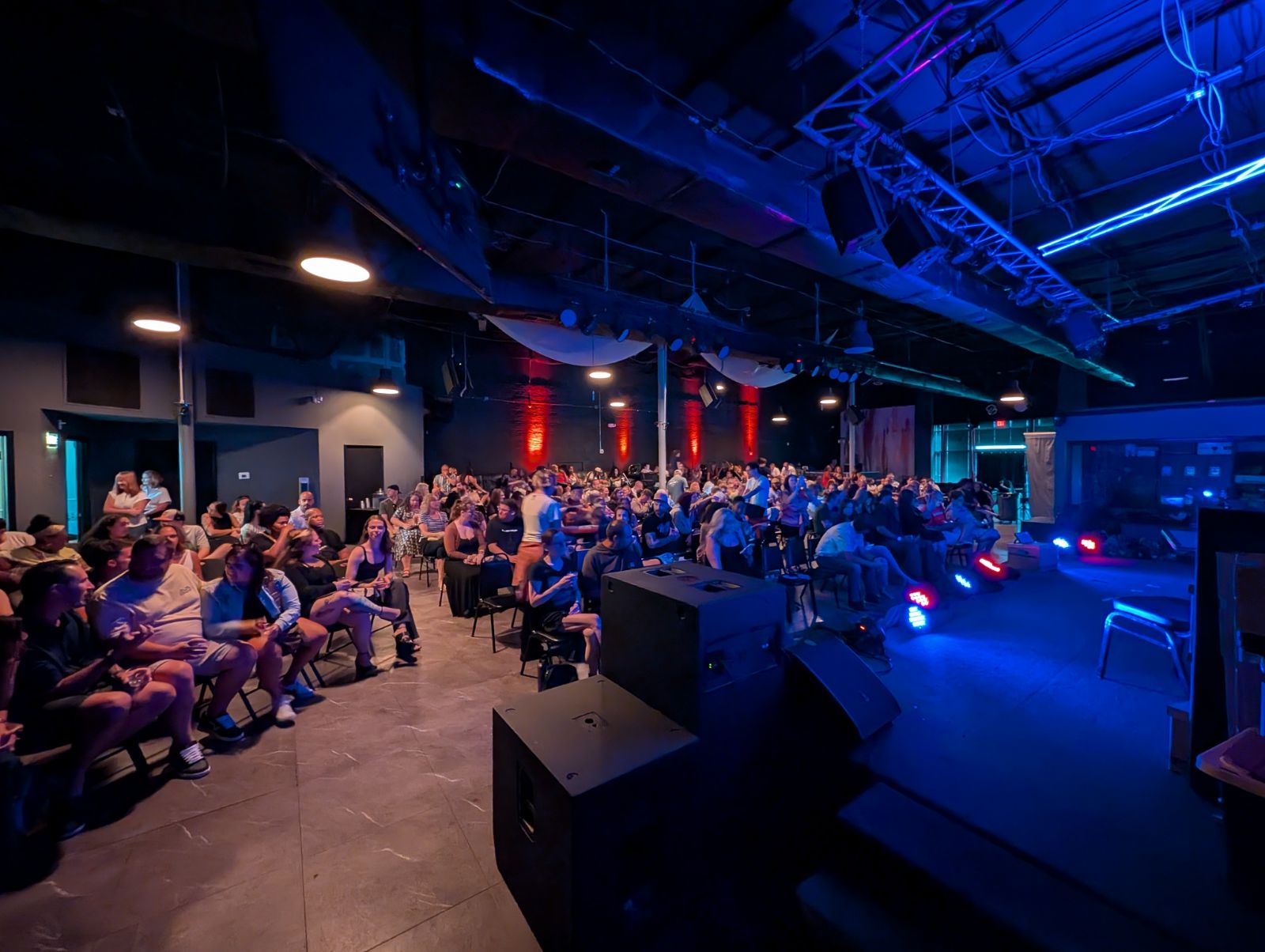 Audience seated in a dark indoor venue with stage lighting, facing a stage area with equipment and blue lighting.