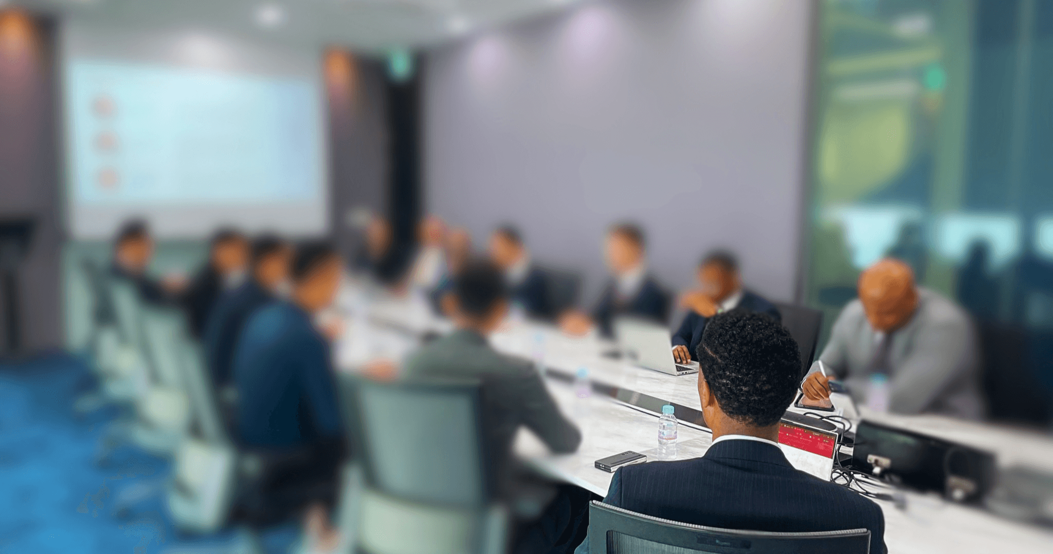 A group of people in a meeting room, seated around a table, with a presentation screen in the background.