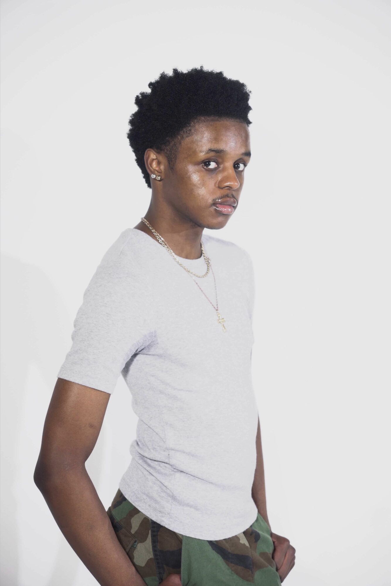 Young woman with short curly hair wearing a light gray T-shirt and camouflage pants, standing against a plain white background.