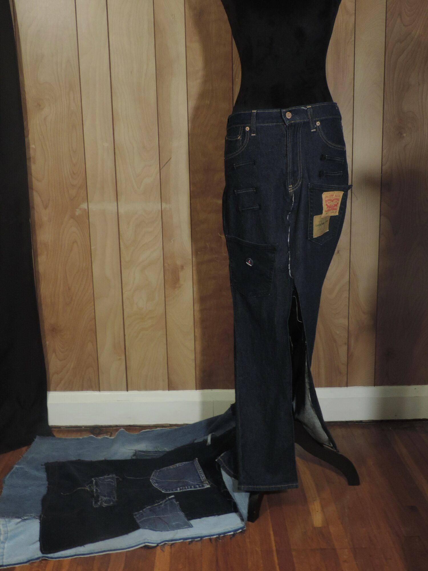 Black mannequin wearing dark jeans with patches, standing on a wooden floor against a wood-paneled wall.