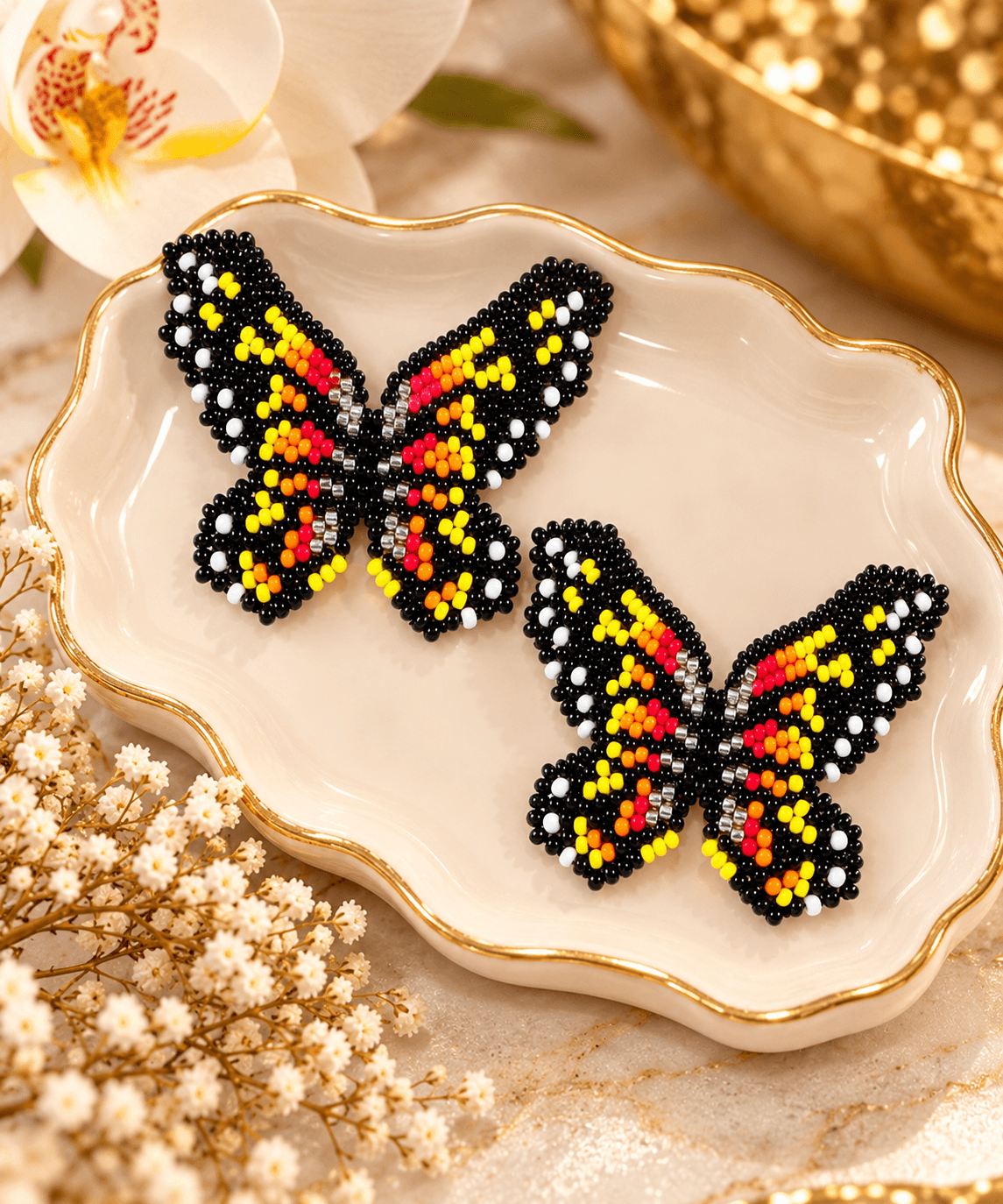 Two beaded butterfly brooches with black, yellow, and red beads on a white dish, surrounded by flowers and decorative items.