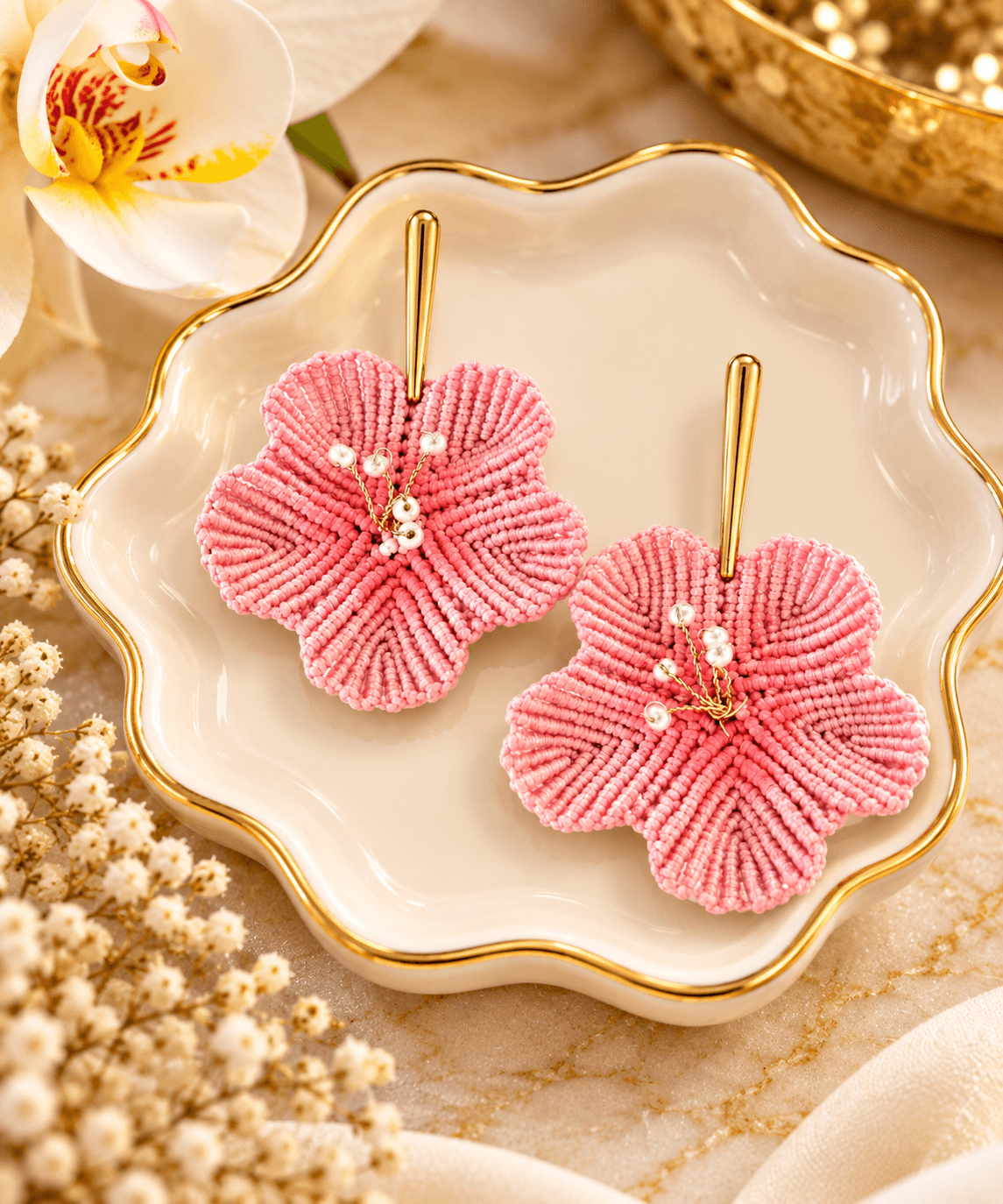 Pink fabric flower-shaped earrings with pearl accents on a white dish, surrounded by flowers and jewelry, in a decorative setting.