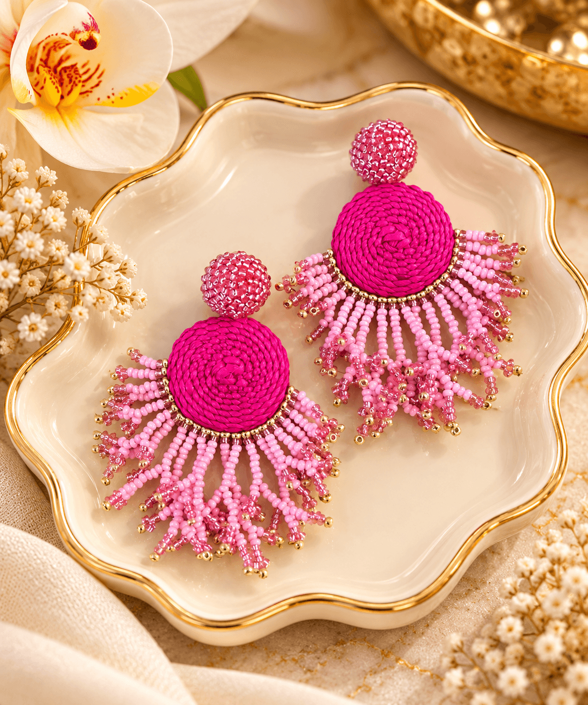 Pink beaded earrings with circular top and fringe hanging below, placed on a decorative dish with flowers nearby.