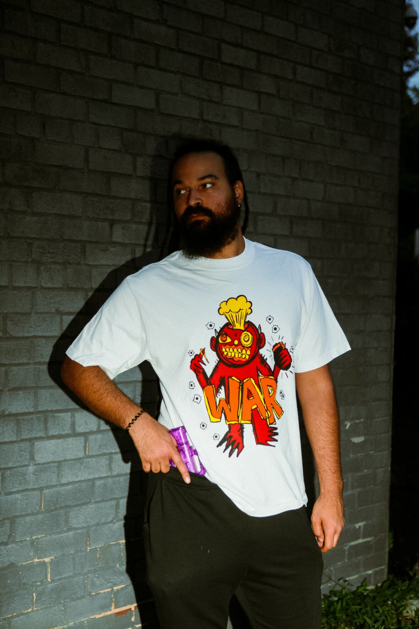Man with a beard wearing a white T-shirt with a colorful cartoon character and text, standing against a brick wall.