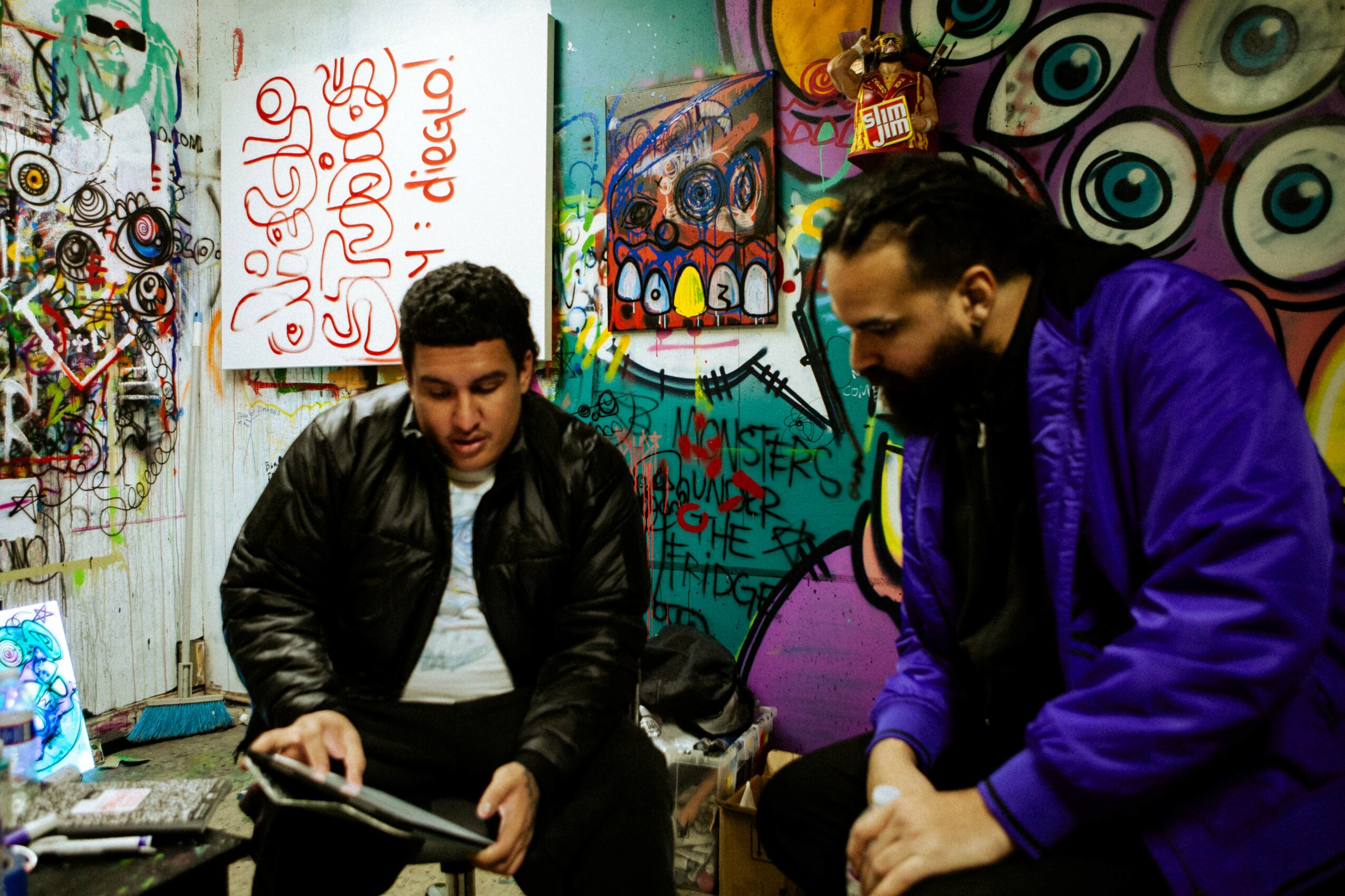 Two men sit in front of colorful graffiti and artwork, one holding a tablet, in an urban art space.