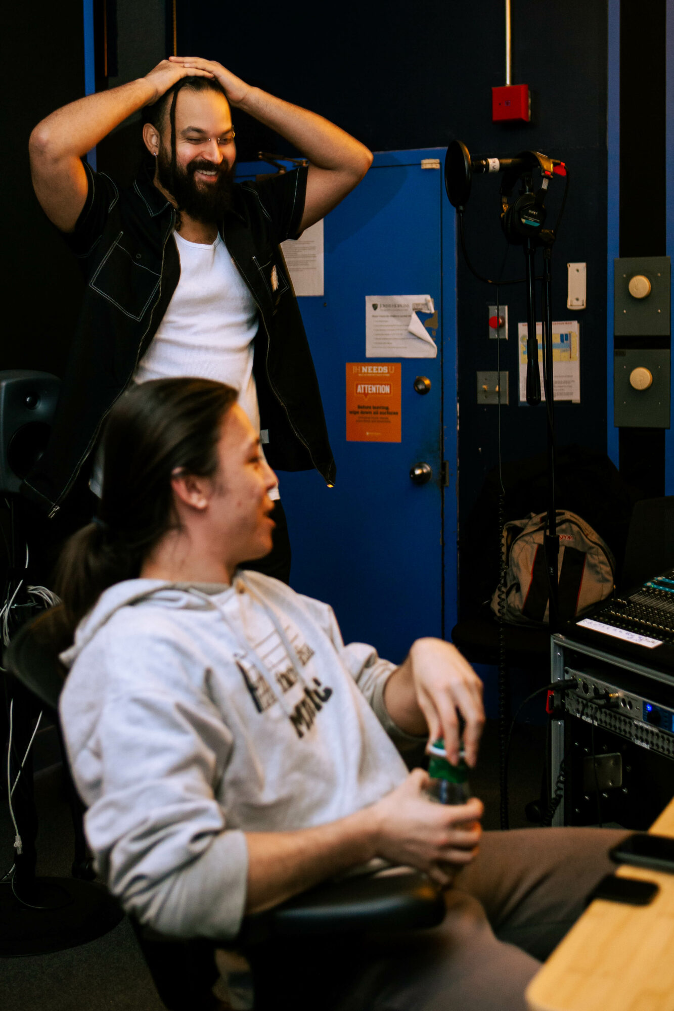 Two people in a recording studio, one standing with hands on head, the other sitting with a drink, equipment visible.