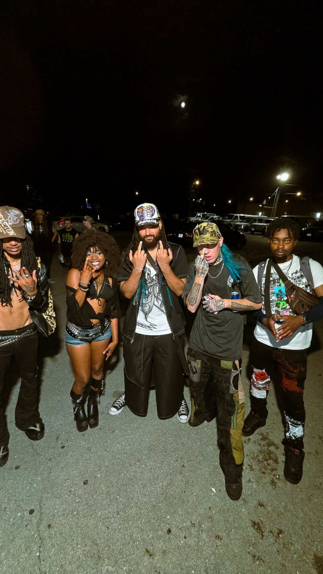 Group of five people standing outdoors at night, some wearing hats and casual clothing, with streetlights in the background.