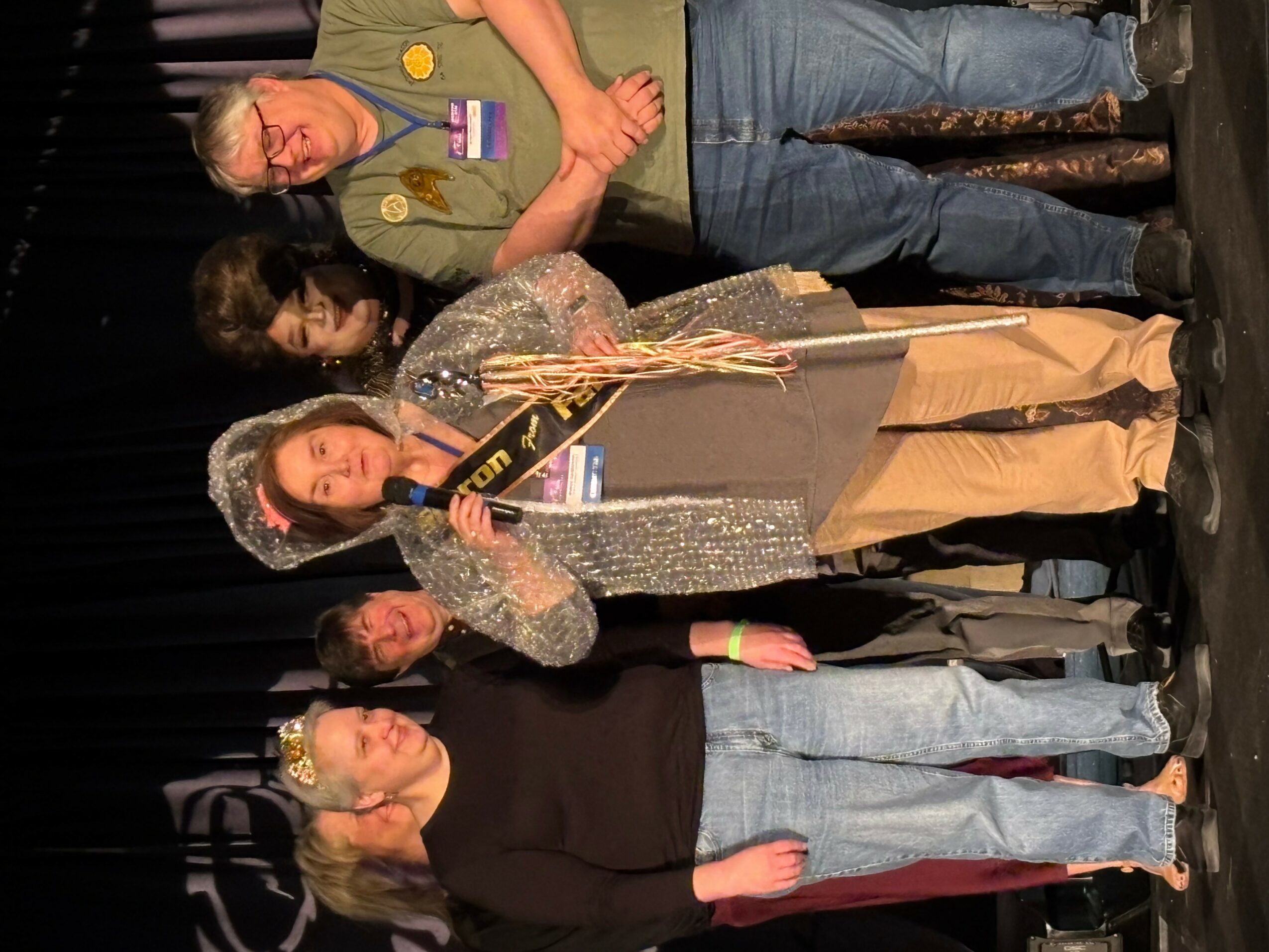 Group of people standing on stage, one woman holding a bouquet of flowers, others watching, black curtain background.